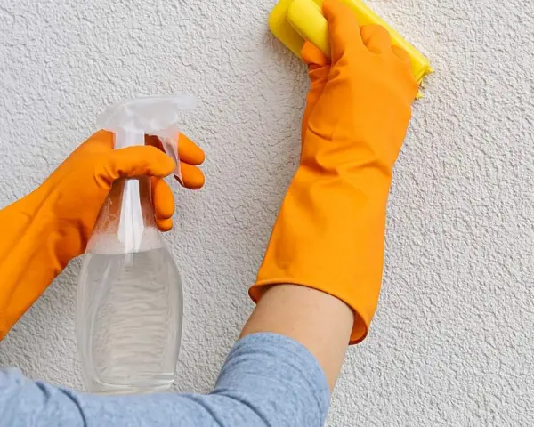 Mani con guanti arancioni puliscono un muro esterno con cappotto, usando uno spray e una spugna gialla.
