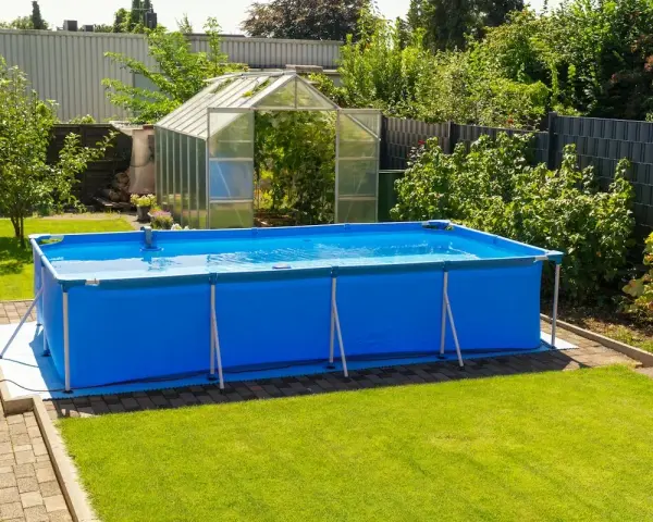 Ampia piscina blu montata in giardino, con serra sullo sfondo. Sembra una piscina senza permessi, pronta per l'estate.