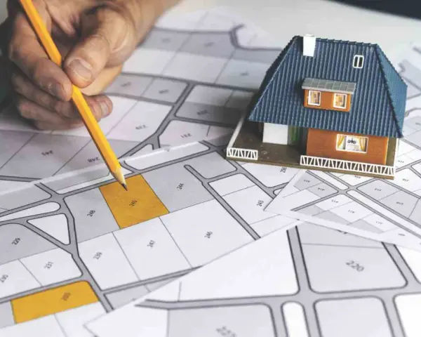 Mano con matita su mappa catastale, con modellino di casa. Pianificazione immobiliare, categoria catastale D10.