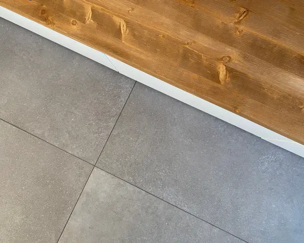 Dettaglio di piastrelle grigie posate con precisione, pronte per attaccare piastrelle al muro con silicone. Sopra, una trave di legno grezzo.