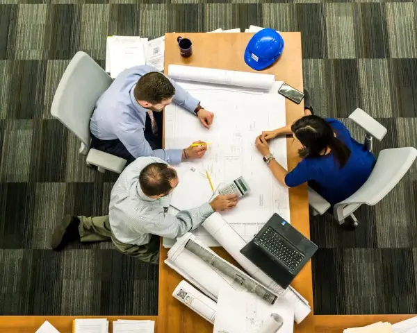 Team di architetti discute progetti su un tavolo, con rotoli di disegni e un casco blu. L'ufficio, con moquette a righe, è ideale per una **categoria catastale A10**.