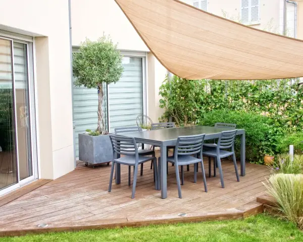 Un tavolo con sedie su un pavimento da giardino su terra, con un ulivo in vaso e una vela parasole.