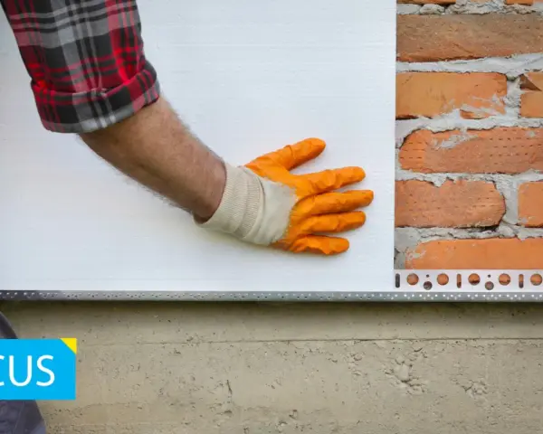 Installazione battiscopa per cappotto esterno: un operaio con guanti arancioni posiziona un pannello isolante bianco su una parete di mattoni, con un profilo metallico alla base.