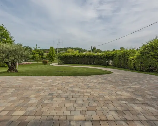 Pavimentazione giardino a secco con blocchi color sabbia, che conducono a un prato verde curato con alberi e siepi.