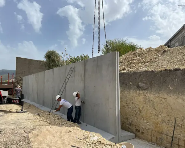 Operai installano un grande muro di contenimento in cemento armato, sollevato da una gru. Il cantiere è in pendenza.