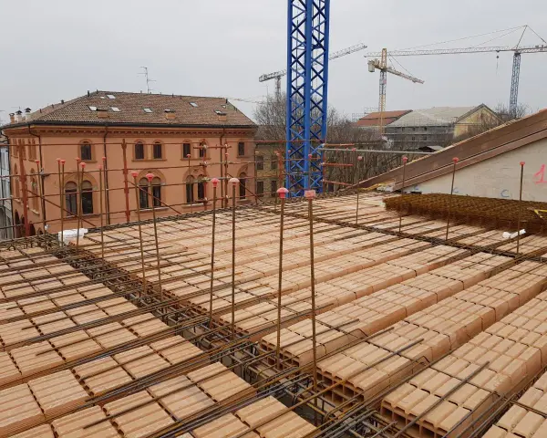 Costruzione di un solaio: mattoni forati e armature in ferro pronti per il getto di cemento. In lontananza, un edificio storico e gru edili.