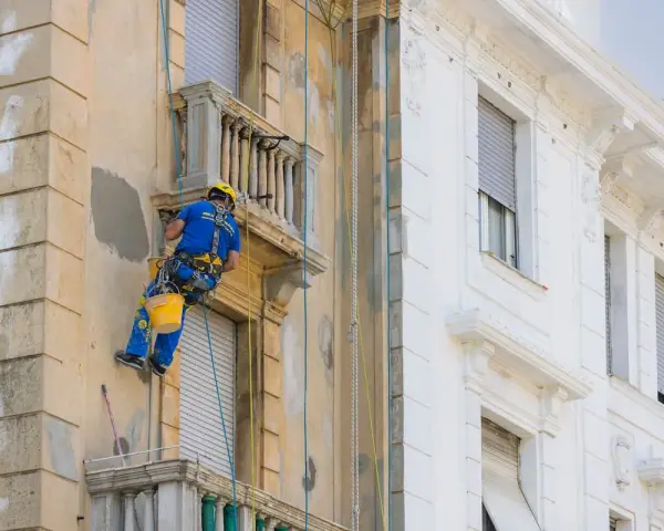 Operaio in corda ripara la facciata di un palazzo, lavorando sui dettagli dei balconi aggettanti.