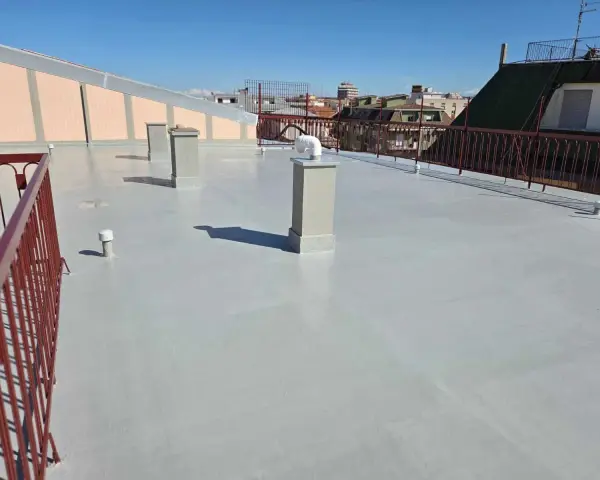 Un tetto in cemento appena impermeabilizzato, con camini e ringhiera rossa, sotto un cielo azzurro.