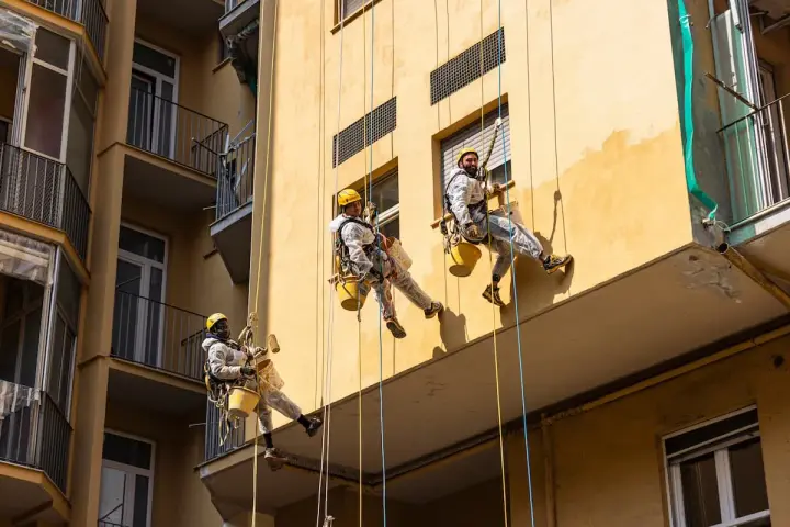 Operai in corda applicano diversi tipi di intonaco per esterni su un edificio giallo.