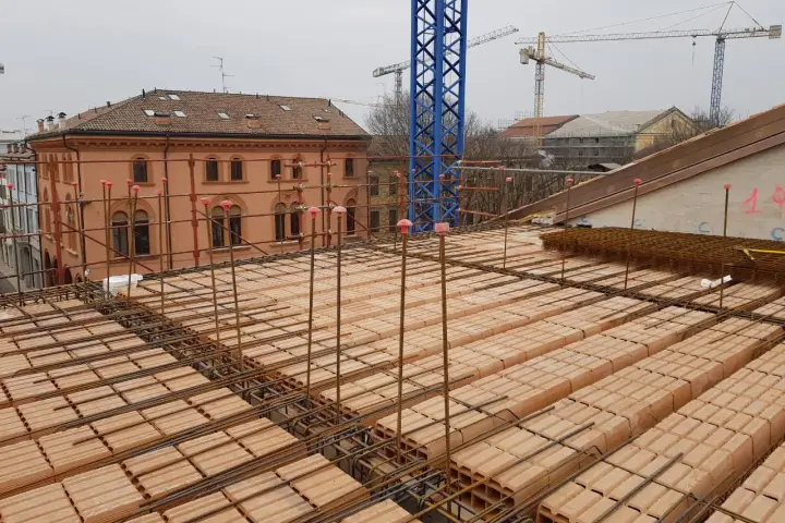 Costruzione di un solaio: mattoni forati e armature in ferro pronti per il getto di cemento. In lontananza, un edificio storico e gru edili.