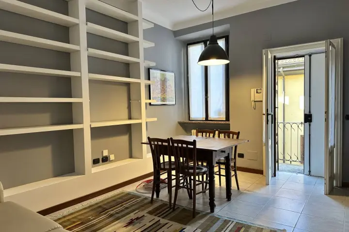 Un soggiorno con libreria a giorno, tavolo in legno e sedie. Dalla porta si intravede un cortile, tipico delle case di ringhiera.