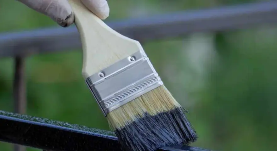Mano con guanto bianco applica vernice scura su ringhiera metallica con pennello, un modo rapido per verniciare ringhiera senza carteggiare.