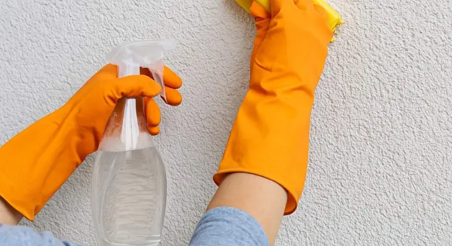 Mani con guanti arancioni puliscono un muro esterno con cappotto, usando uno spray e una spugna gialla.