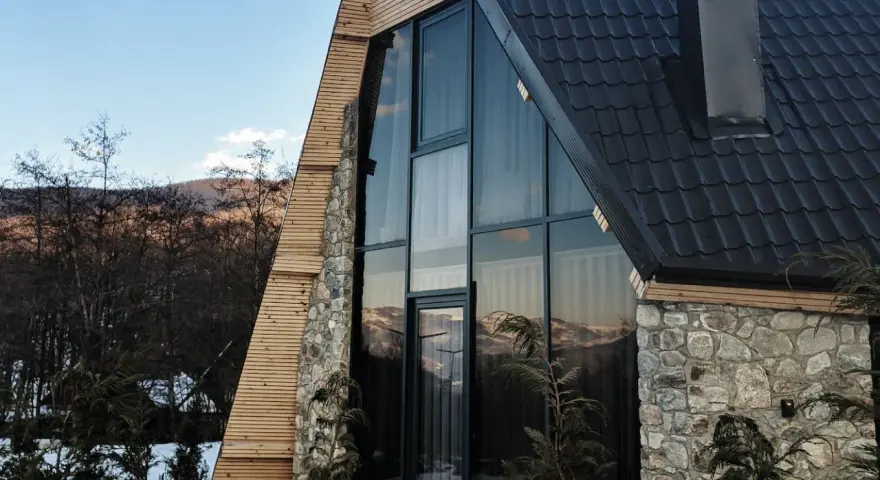 Moderna ristrutturazione casa di montagna: tetto scuro, ampie vetrate con vista sui monti, pietra e legno.