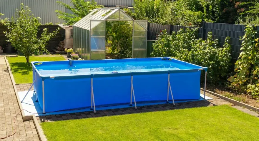 Ampia piscina blu montata in giardino, con serra sullo sfondo. Sembra una piscina senza permessi, pronta per l'estate.