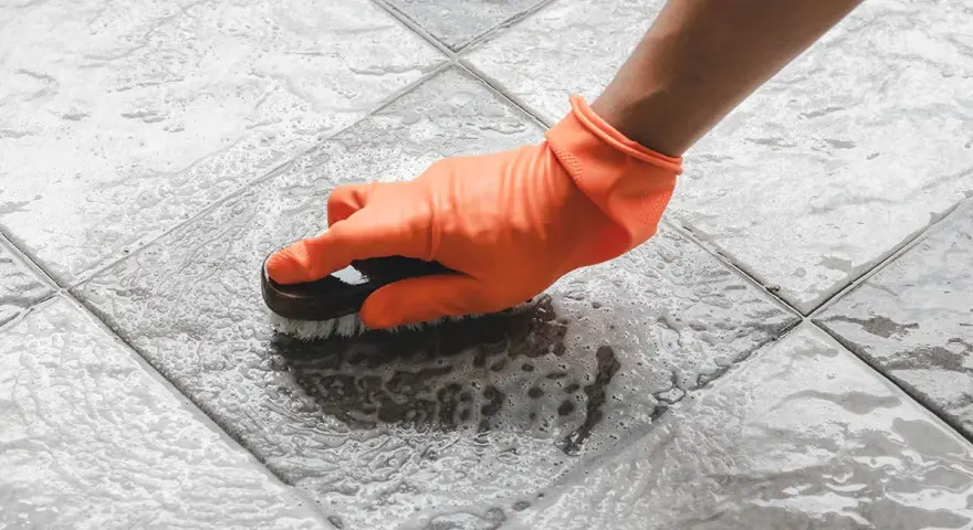Mano con guanto arancione che spazzola un pavimento piastrellato bagnato, mostrando come togliere ruggine dal pavimento con schiuma.