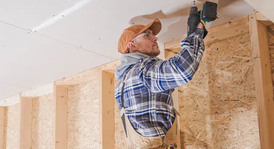 Uomo con trapano installa pannelli per isolamento acustico fai da te sul soffitto di una stanza in costruzione.