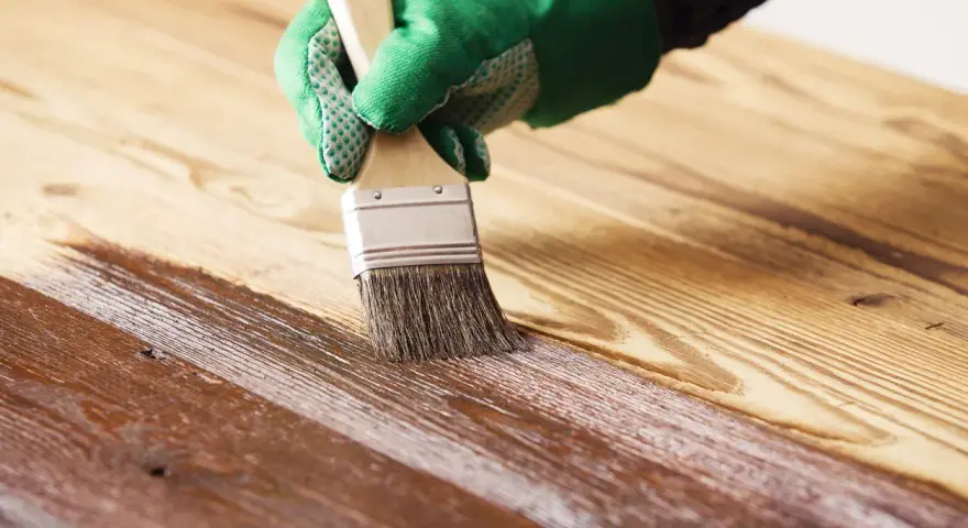 Mano guantata applica vernice protettiva con pennello per impermeabilizzare il legno, creando una finitura lucida.