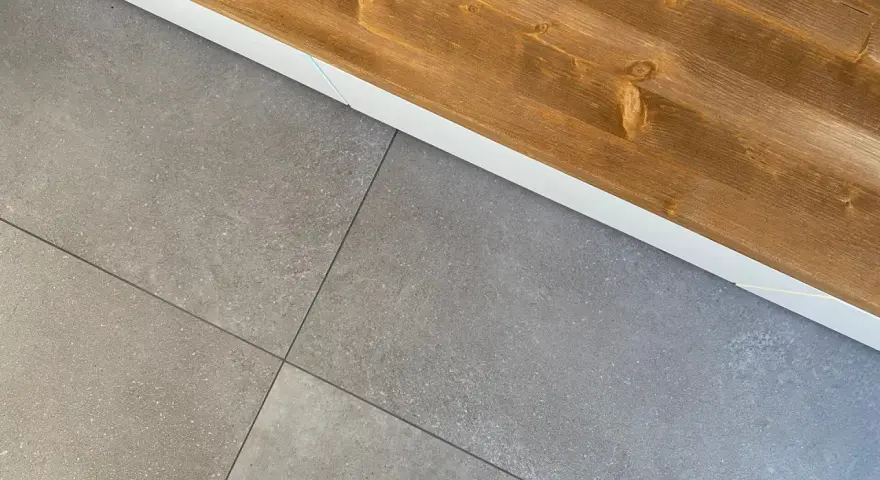 Dettaglio di piastrelle grigie posate con precisione, pronte per attaccare piastrelle al muro con silicone. Sopra, una trave di legno grezzo.