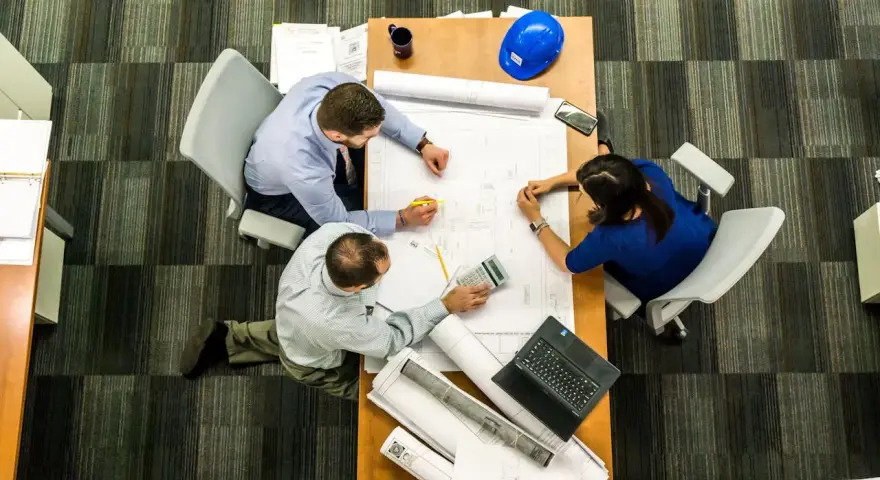 Team di architetti discute progetti su un tavolo, con rotoli di disegni e un casco blu. L'ufficio, con moquette a righe, è ideale per una **categoria catastale A10**.