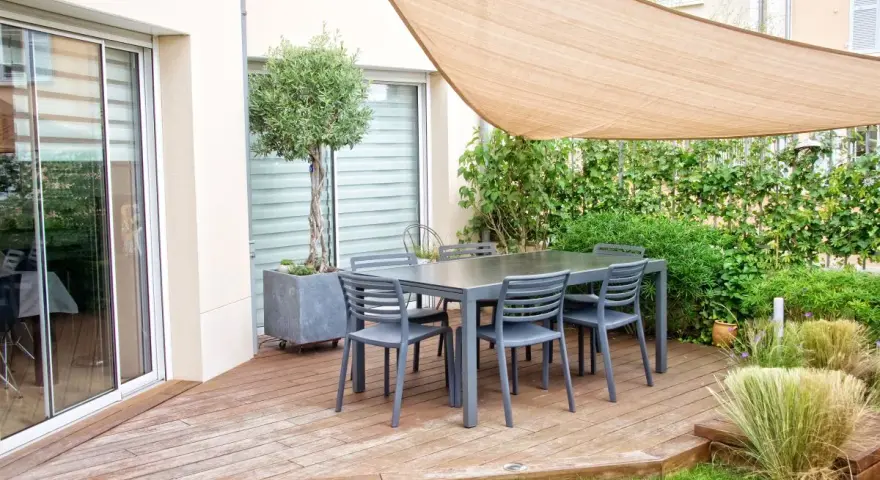 Un tavolo con sedie su un pavimento da giardino su terra, con un ulivo in vaso e una vela parasole.