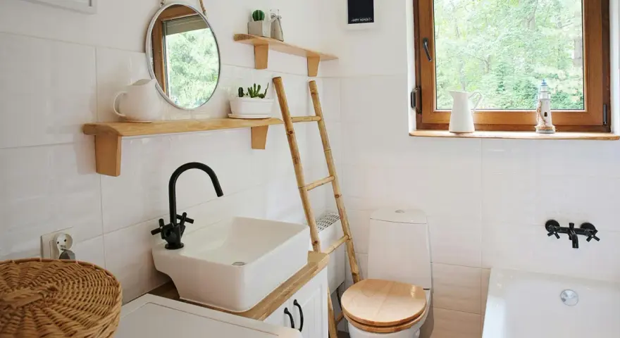 Bagno piccolo con piastrelle bianche lucide, lavabo squadrato, specchio rotondo e scala in bambù. Idee piastrelle bagno piccolo: la luminosità è la chiave.