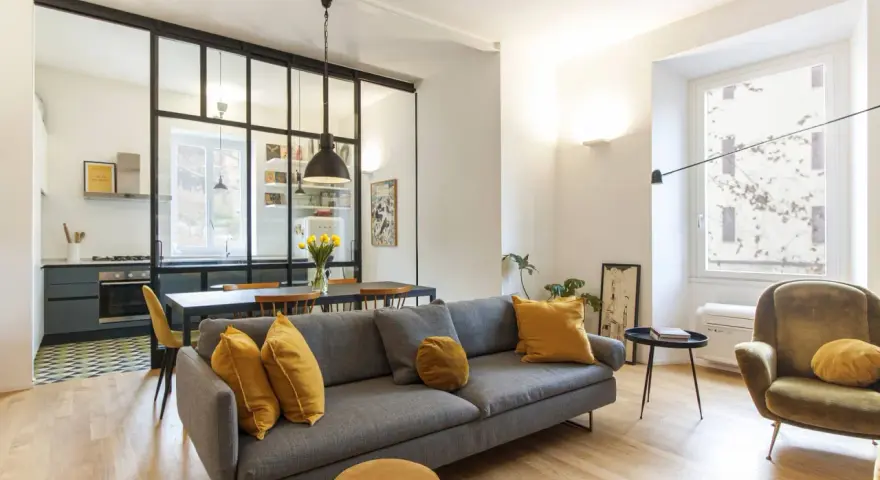 Ampia zona giorno con divano grigio e poltrona verde, separata dalla cucina da una moderna mezza parete divisoria cucina soggiorno in ferro e vetro.