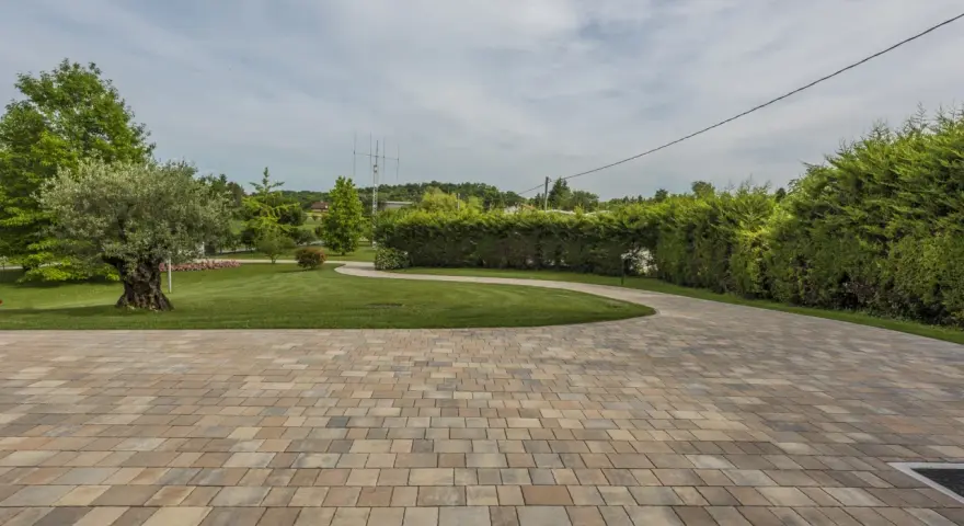 Pavimentazione giardino a secco con blocchi color sabbia, che conducono a un prato verde curato con alberi e siepi.