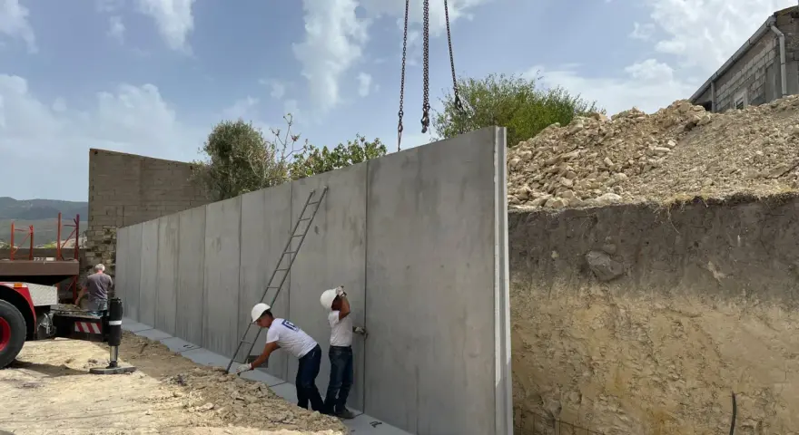Operai installano un grande muro di contenimento in cemento armato, sollevato da una gru. Il cantiere è in pendenza.