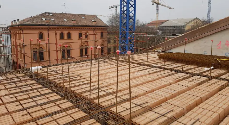 Costruzione di un solaio: mattoni forati e armature in ferro pronti per il getto di cemento. In lontananza, un edificio storico e gru edili.