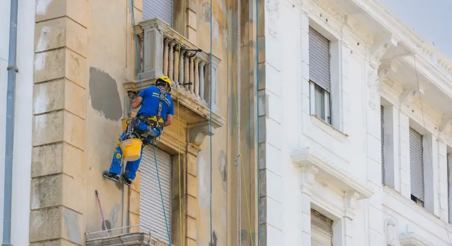 Operaio in corda ripara la facciata di un palazzo, lavorando sui dettagli dei balconi aggettanti.