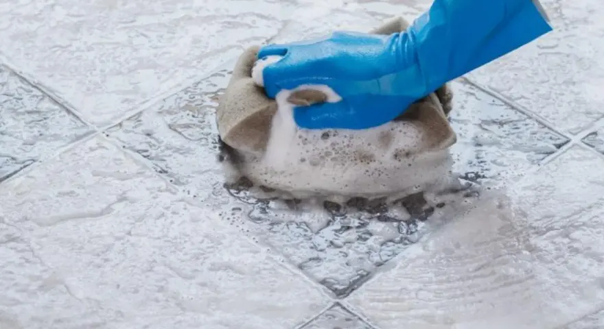 Mano con guanto blu che pulisce ceramica macchiata con spugna e schiuma.