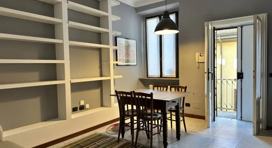 Un soggiorno con libreria a giorno, tavolo in legno e sedie. Dalla porta si intravede un cortile, tipico delle case di ringhiera.