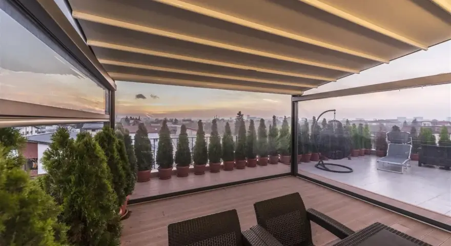Terrazzo trasformato in stanza con chiusura trasparente. Arredi da esterno, piante e vista panoramica al tramonto.