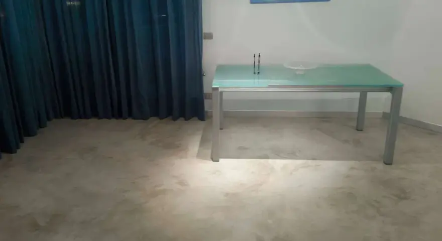 Un tavolo moderno con piano in vetro poggia su un **pavimento in cemento grezzo** dall'aspetto materico, con tende blu sullo sfondo.