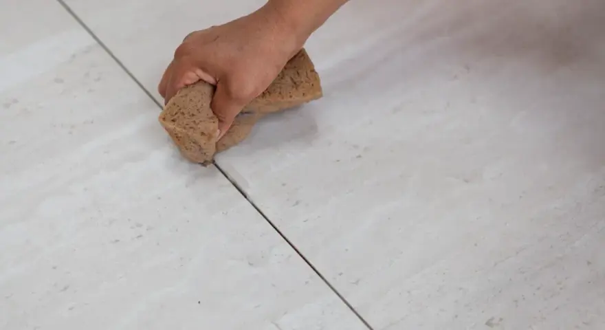 Mano che stuccare piastrelle con una spugna, rifinendo le fughe di un pavimento chiaro.