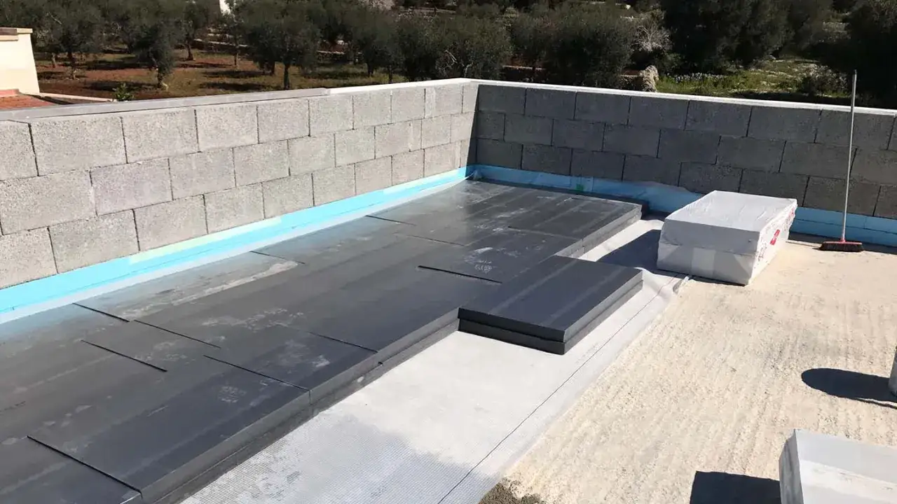 Posa di pannelli per isolante per terrazzi su una struttura in blocchi di cemento, con rotoli di materiale protettivo e una scopa in attesa.