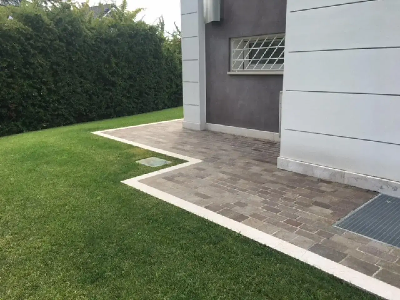 Un moderno marciapiede in pietra circonda la casa, con un prato verde e una siepe. Ottime idee marciapiede casa per un design elegante.