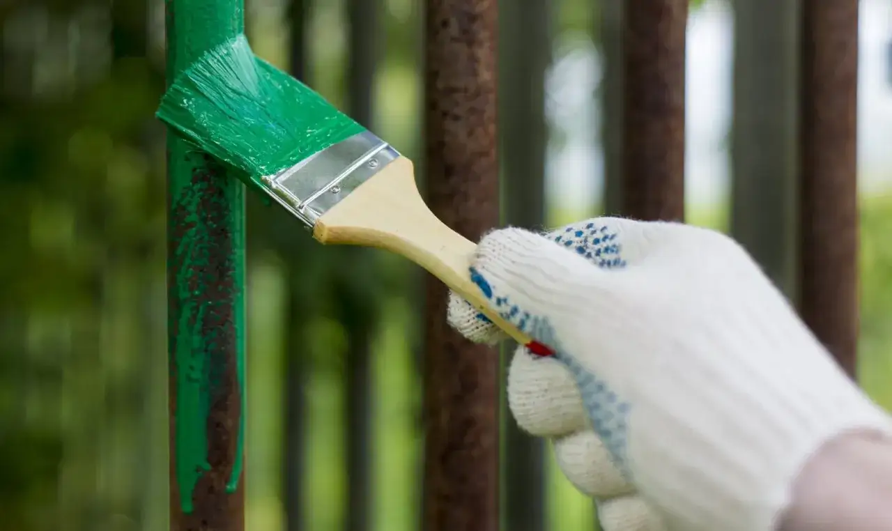 Mano guantata applica vernice verde su ringhiera arrugginita, un modo rapido per verniciare ringhiera senza carteggiare.