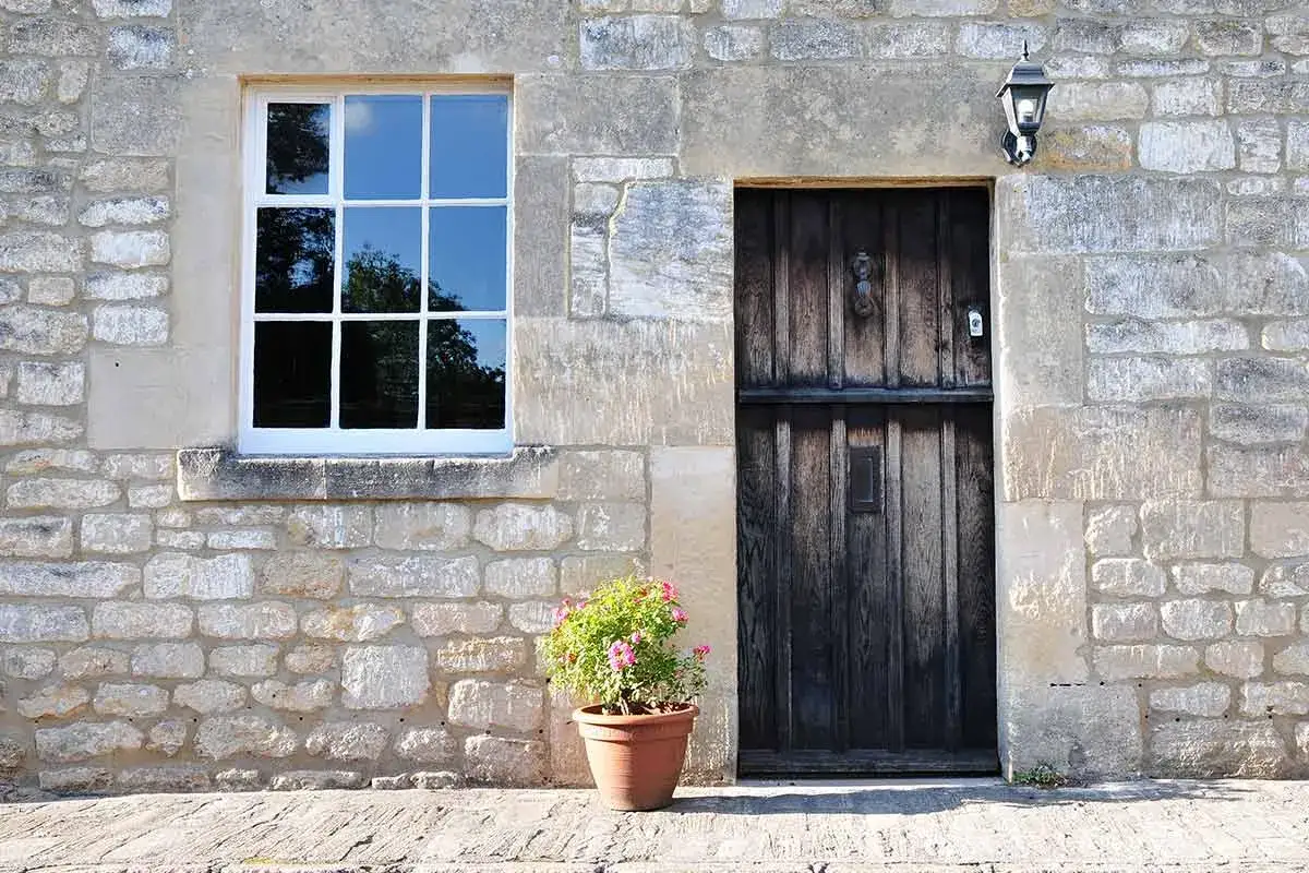 Facciate in pietra e mattoni grezzi, con una porta in legno scuro e una finestra bianca. Un vaso di fiori rosa aggiunge un tocco di colore.