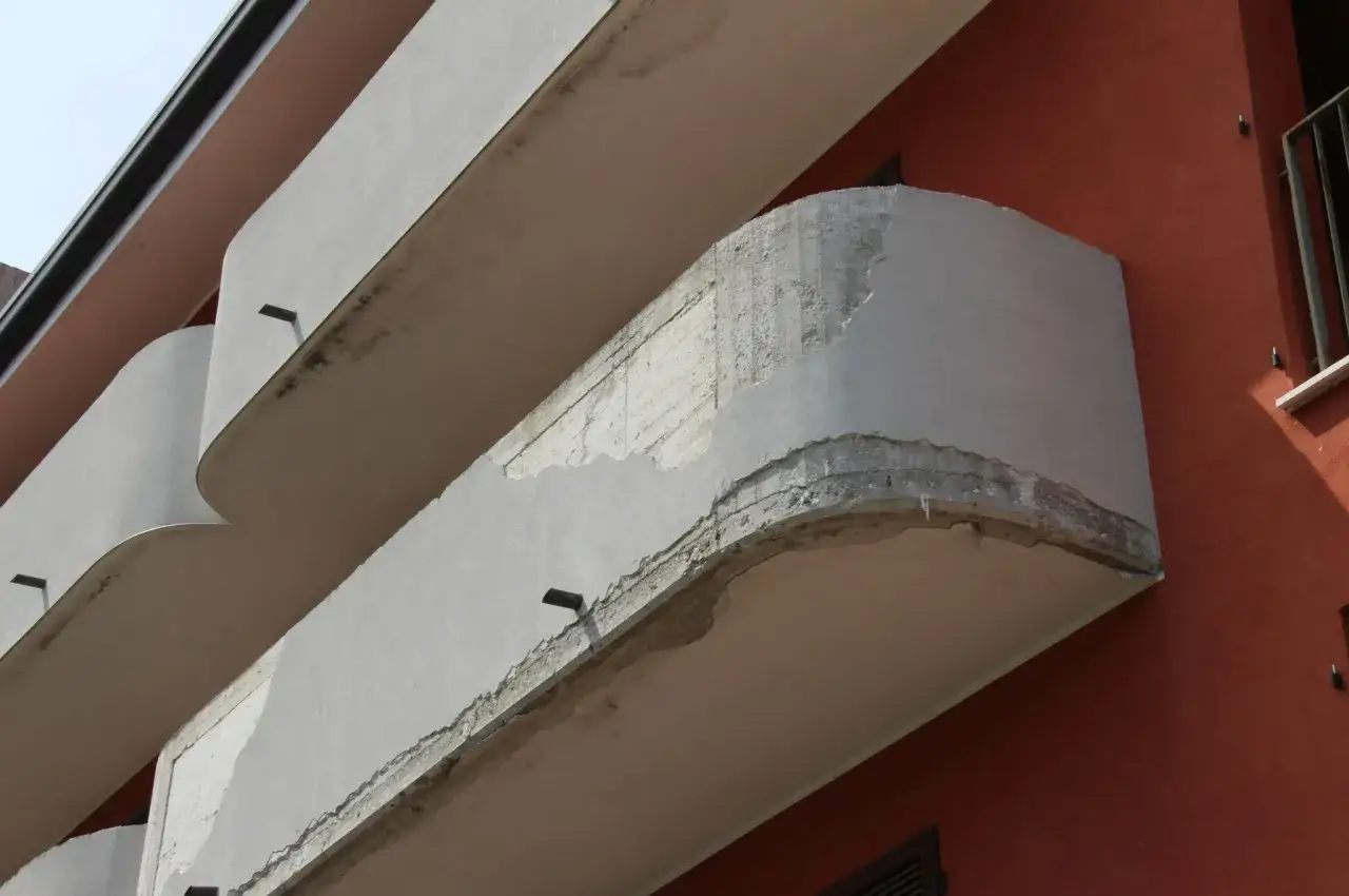 Dettaglio di balconi aggettanti di un edificio con intonaco scrostato che rivela il cemento armato sottostante.