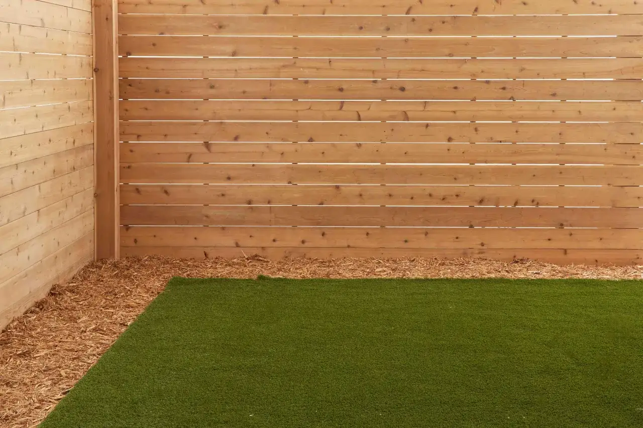 Angolo di un giardino con prato sintetico e pacciamatura di legno, delimitato da una moderna recinzione in listelli orizzontali, ideale per barriere antirumore fai da te.