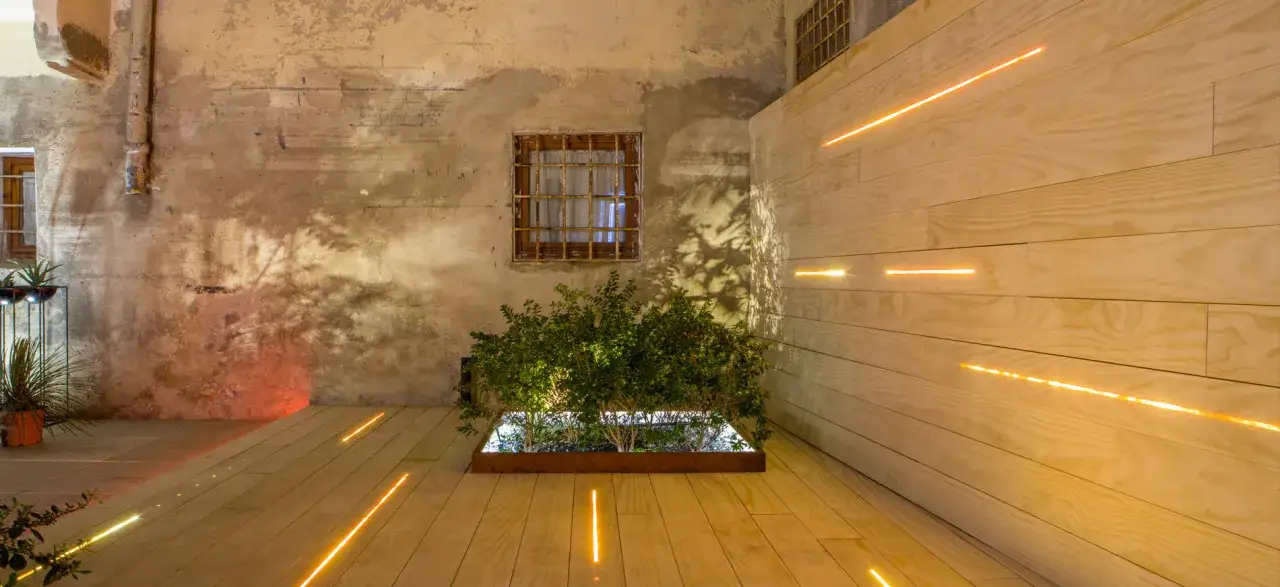 Un giardino moderno con un muretto in legno illuminato da luci soffuse. Un'aiuola centrale con piante verdi crea un punto focale.