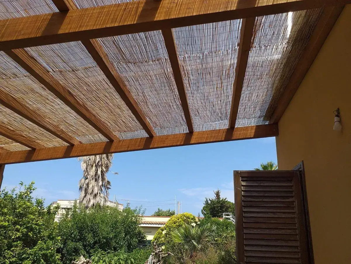 Tettoia fai da te in canne e legno, con vista su un giardino rigoglioso e cielo azzurro.
