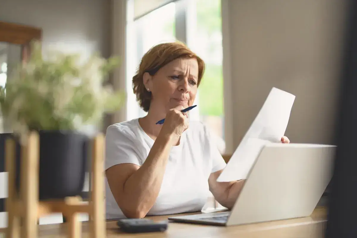 Donna pensierosa con fogli e laptop, forse sta calcolando chi paga IMU.